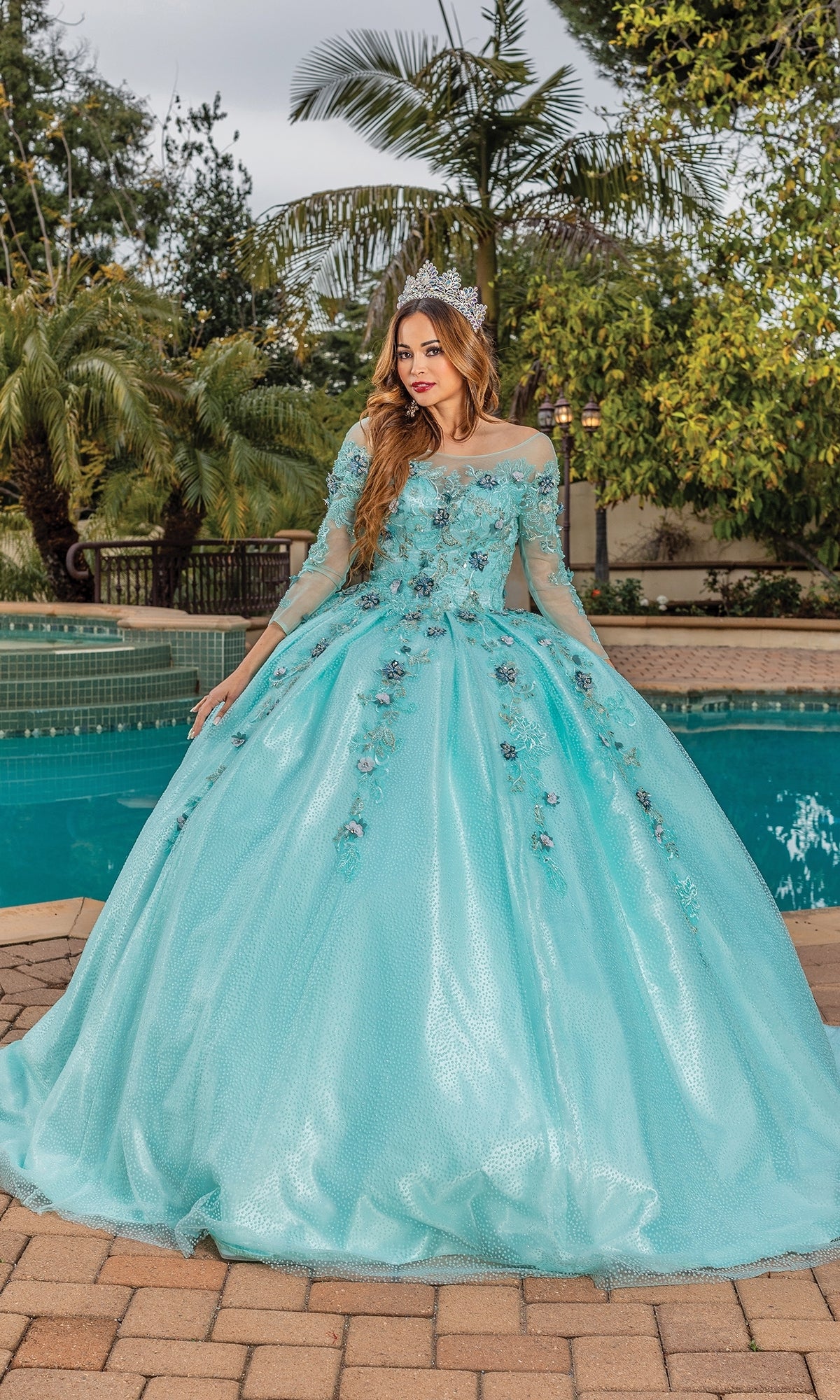 A woman stands by a pool in the Quinceanera Dress 1847 By Dancing Queen, a mint green gown with floral embellishments and a tiara, surrounded by palm trees and greenery.